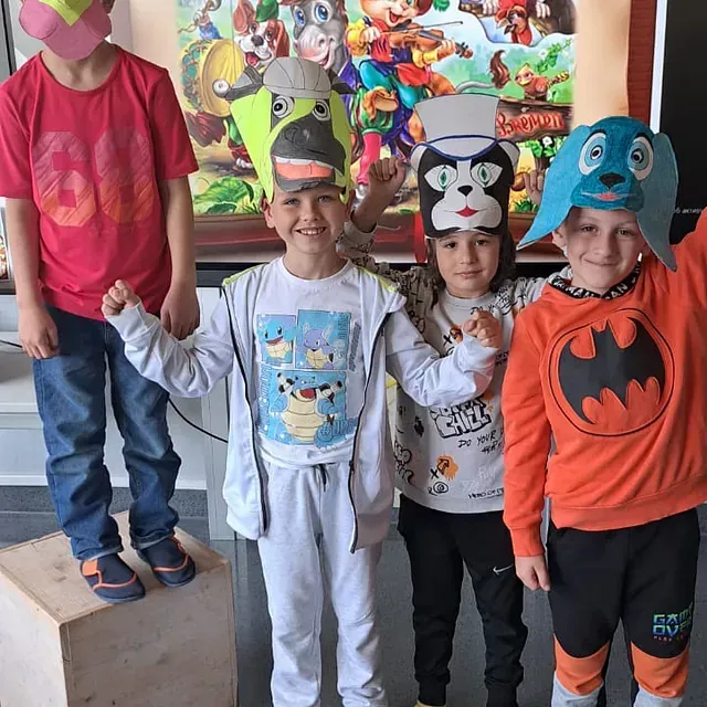 Kinder mit Tiermasken posieren vor einem farbenfrohen Wandgemälde, einige heben die Arme.