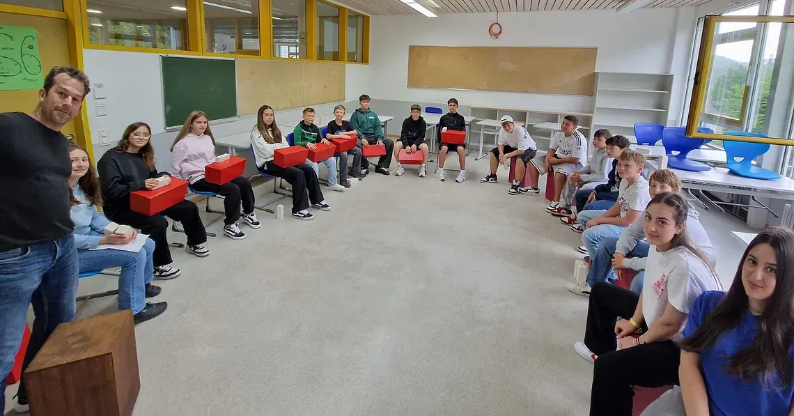 Schüler und ein Mann stehen im Kreis und halten rote Kisten in einem Klassenzimmer.