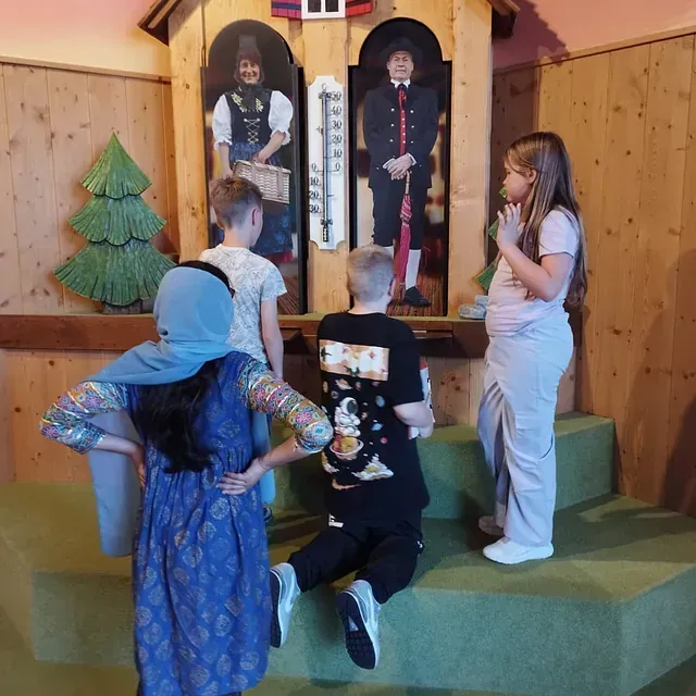 Kinder betrachten eine Kuckucksuhr mit traditionellen Figuren. Grüne Bühne, hölzerner Hintergrund.