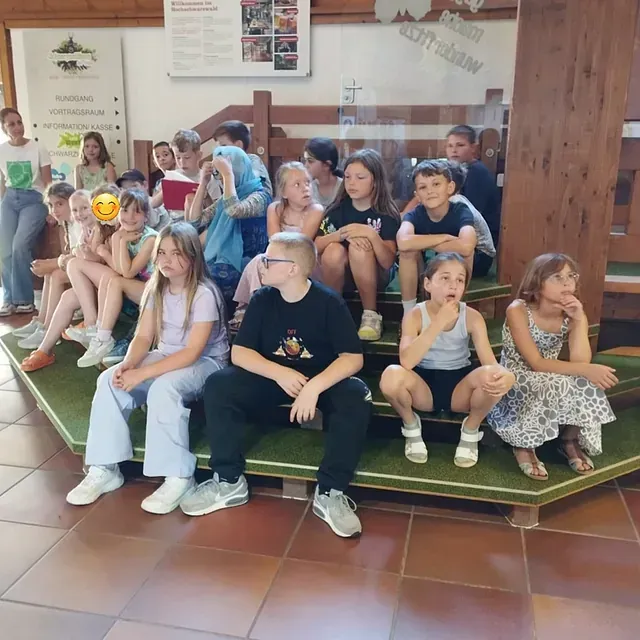 Eine Gruppe Kinder sitzt auf einer Treppe und posiert. Einige schauen in die Kamera, andere woanders hin. Innenaufnahme.