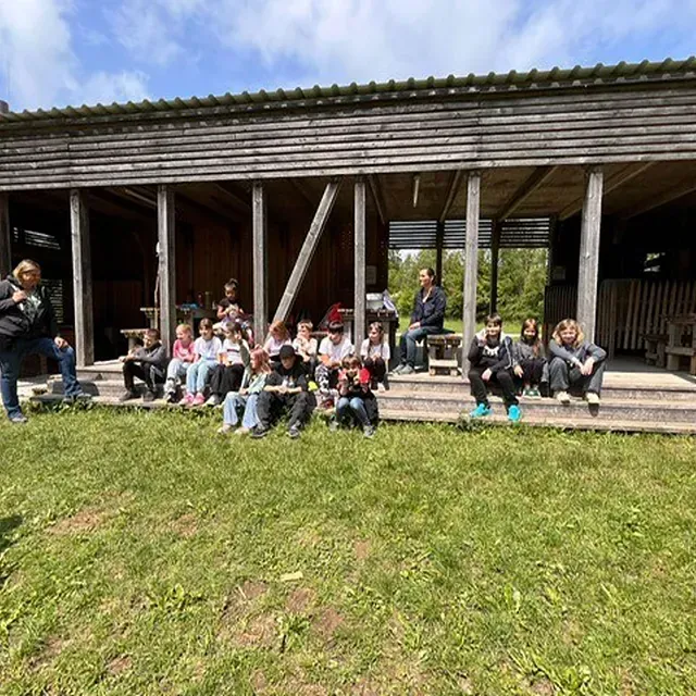 Eine Gruppe Kinder sitzt auf den Stufen eines Holzgebäudes, einige halten Gegenstände in den Händen. Davor befindet sich eine Rasenfläche.