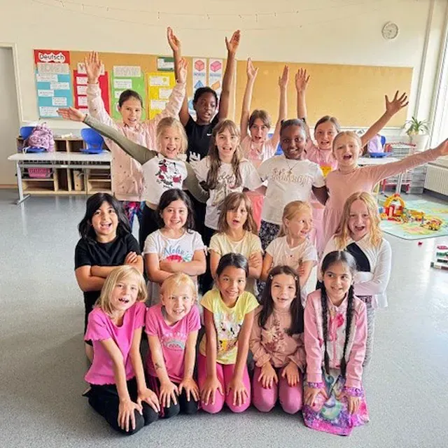 Eine Gruppe Mädchen in einem Klassenzimmer, die Arme in die Luft erhoben, lächelnd. Einige knien, in Rosa gekleidet.