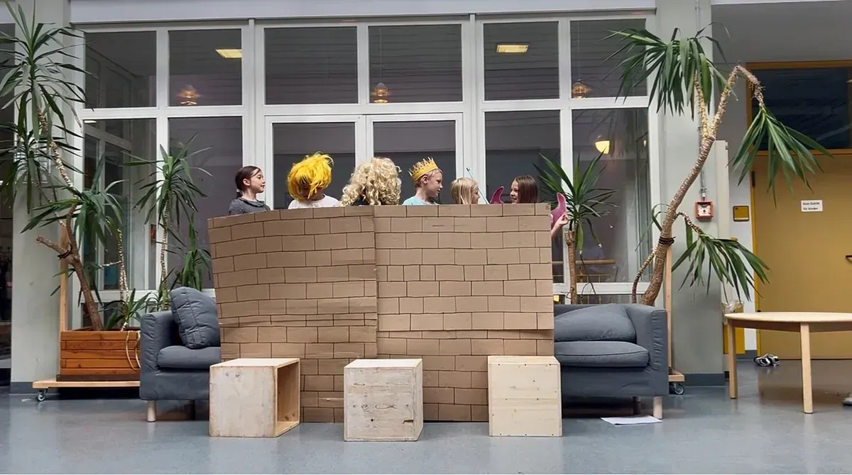 Menschen hinter einer braunen Backsteinmauer; zwei graue Sofas, Holzkisten und Pflanzen in einem großen Raum mit Fenstern.