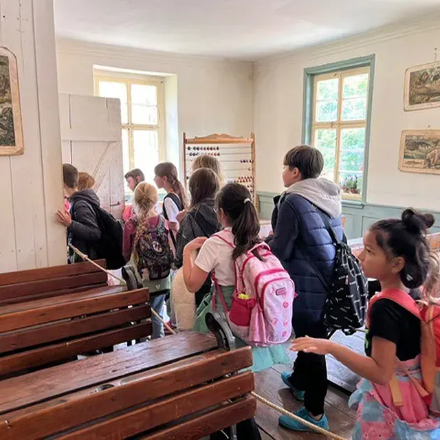 Schulkinder schauen in einen Raum, einige tragen Rucksäcke. Im Vordergrund Holzbänke, im Hintergrund Fenster.