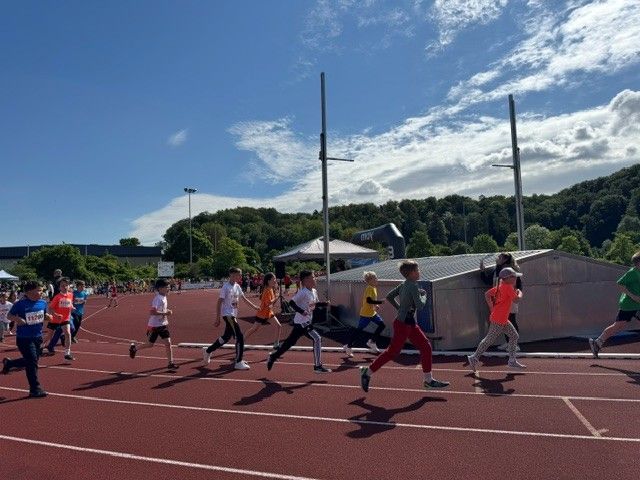 Runners sprint on a track under a sunny sky, with tents and hills in the background.