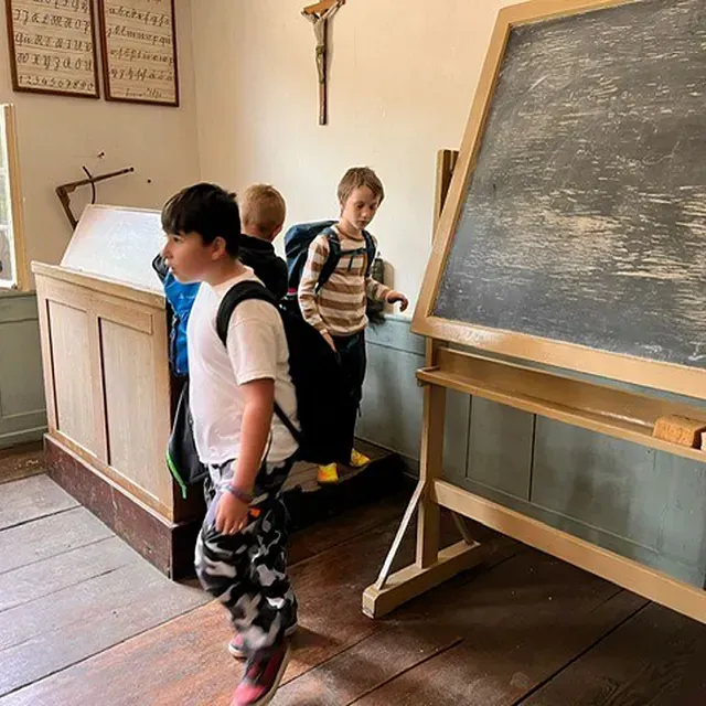 Kinder in einem altmodischen Klassenzimmer mit Tafel und Schreibtisch. Ein Junge trägt eine Tarnhose.
