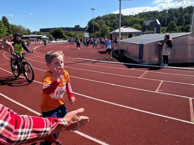 Runner in orange jersey on a red track, reaching toward a spectator’s outstretched hand at a stadium finish area.