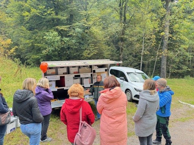 Menschen versammeln sich um einen Lastwagen mit Bienenstöcken; ein Mann spricht, weißes Auto, Wald im Hintergrund.