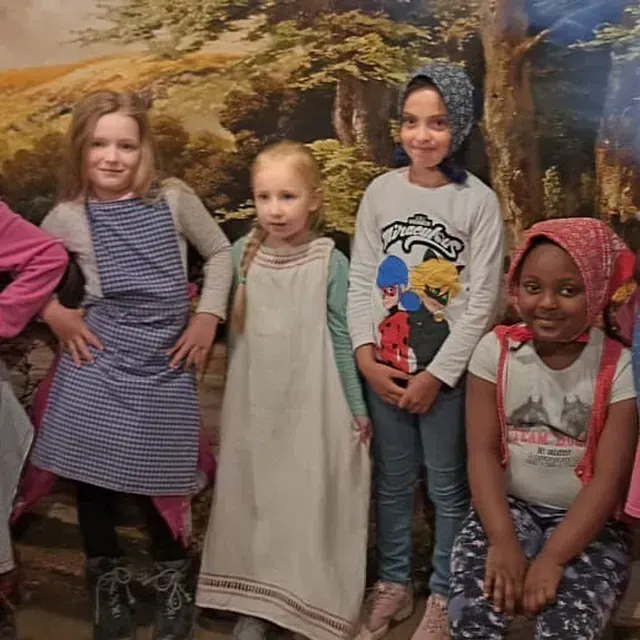 Vier Kinder vor einer bemalten Kulisse. Einige tragen Kopftücher und Schürzen.