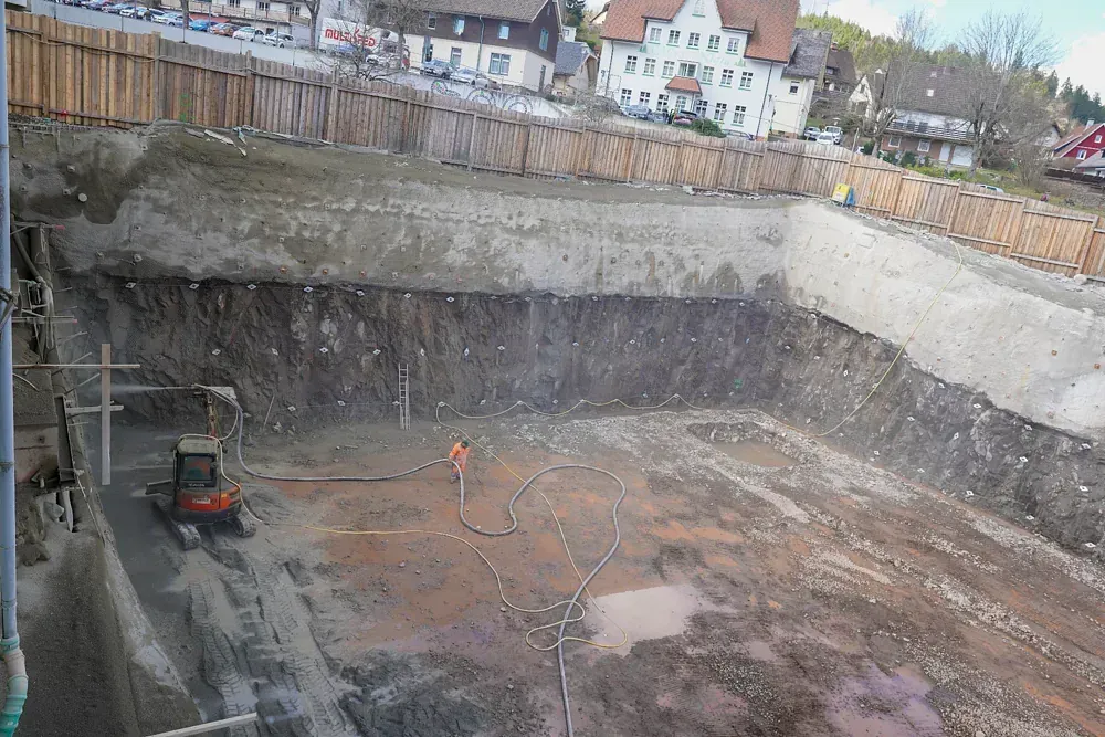 Baustelle: Ausgehobene Baugrube mit Stützmauern, kleiner Bagger, Arbeiter und Gebäude im Hintergrund.