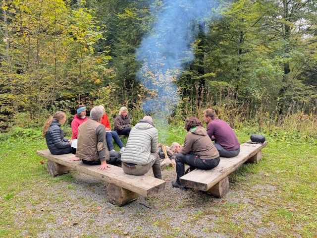 Eine Gruppe sitzt um ein Lagerfeuer in einem Wald, Bänke umgeben die Flammen und den aufsteigenden Rauch.