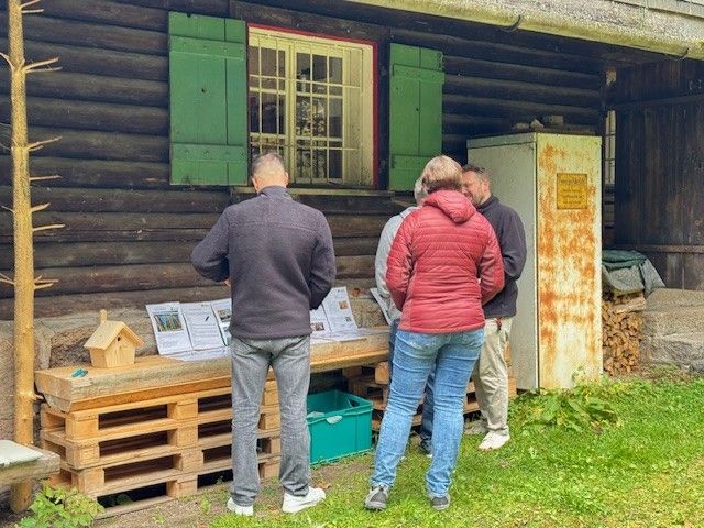 Menschen lesen die Informationstafeln vor einer Blockhütte mit grünen Fensterläden.