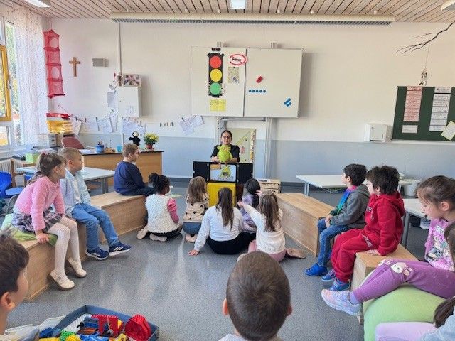 Kinder in einem Klassenzimmer hören sich eine Präsentation zum Thema Verkehrssicherheit an; eine Ampel ist zu sehen.