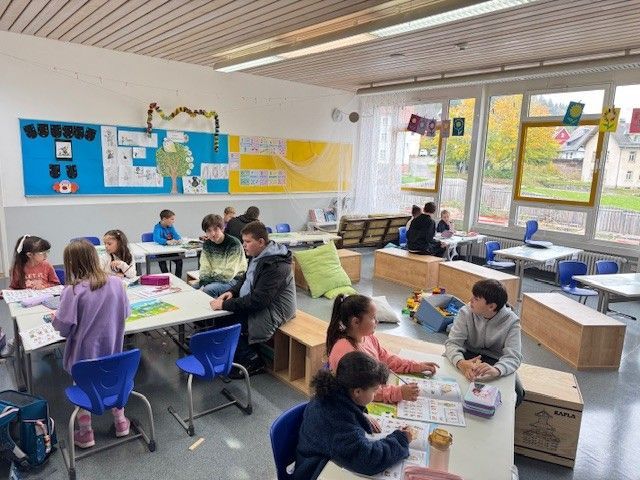 Die Schüler arbeiten an Tischen in einem Klassenzimmer. Durch die Fenster sieht man das Herbstlaub. Einige Schüler sitzen auf Holzkonstruktionen.
