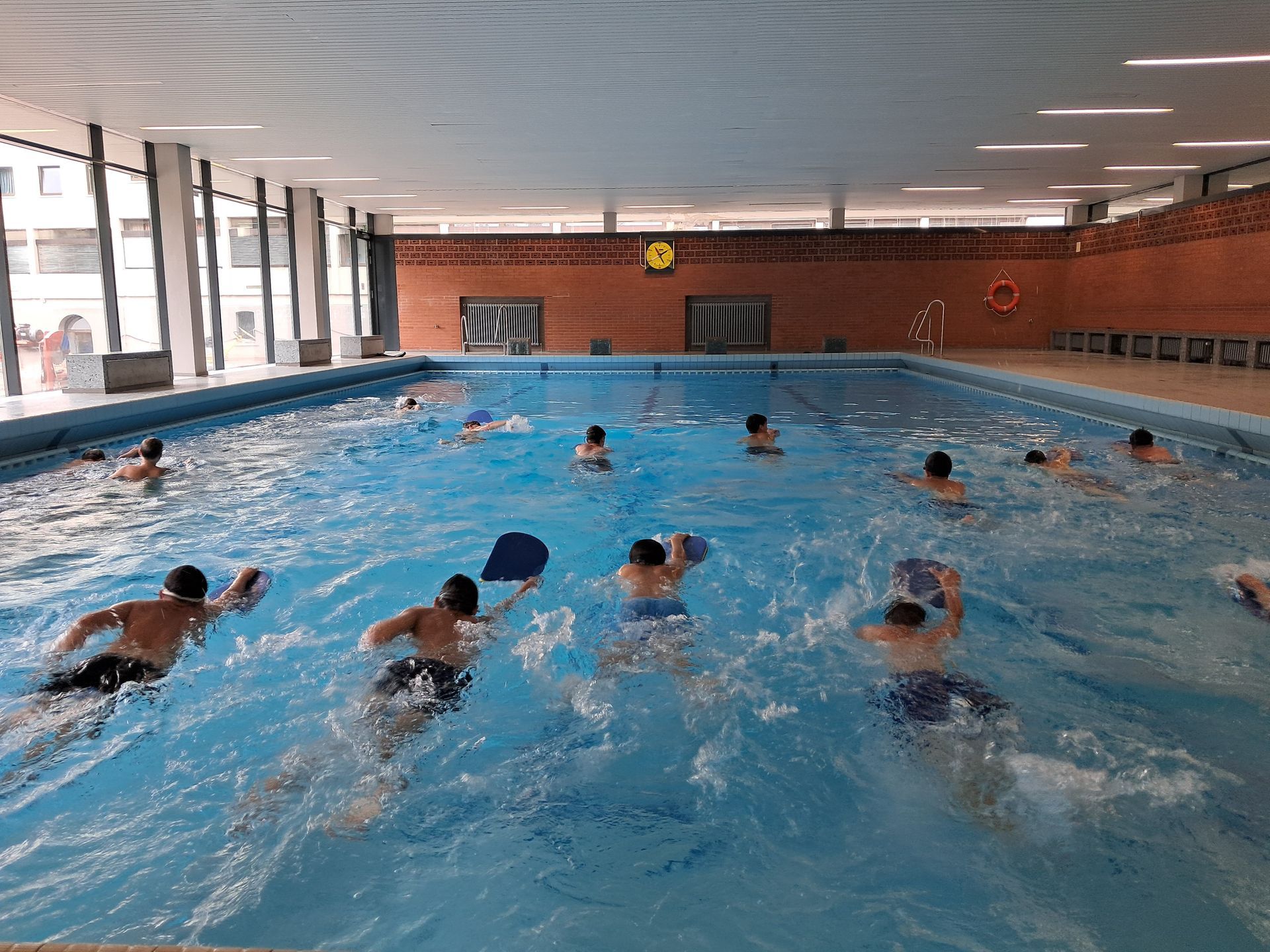 Menschen schwimmen in einem großen Hallenbad, einige mit Schwimmbrettern.