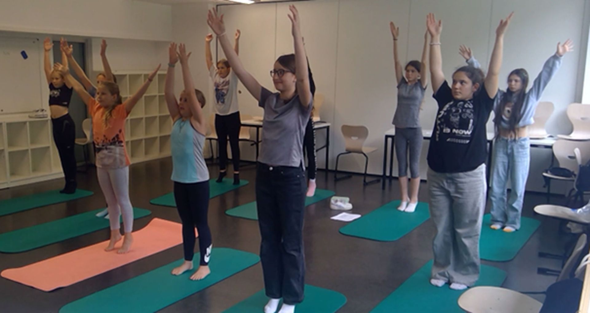Eine Gruppe von Menschen macht Yoga in einem Klassenzimmer; sie strecken ihre Arme nach oben, während sie auf Yogamatten stehen.