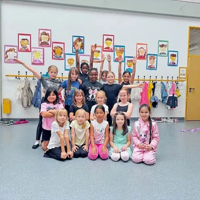 Eine Gruppe Kinder posiert in einem Tanzstudio vor einer Wand mit Porträts. Einige knien, andere stehen und heben die Hände.