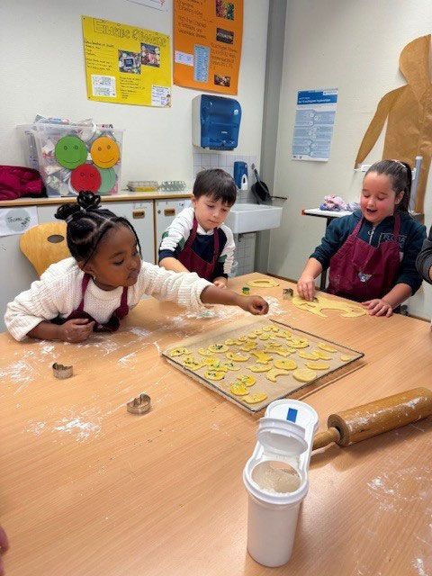 Drei Kinder in Schürzen backen an einem Tisch Plätzchen mit Ausstechformen und einem Nudelholz.