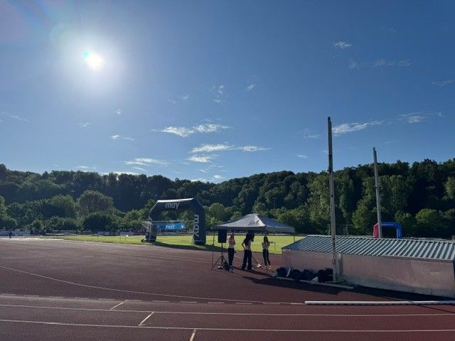 Sunlit outdoor track and field with a few people near tents and a wooded hillside in the background