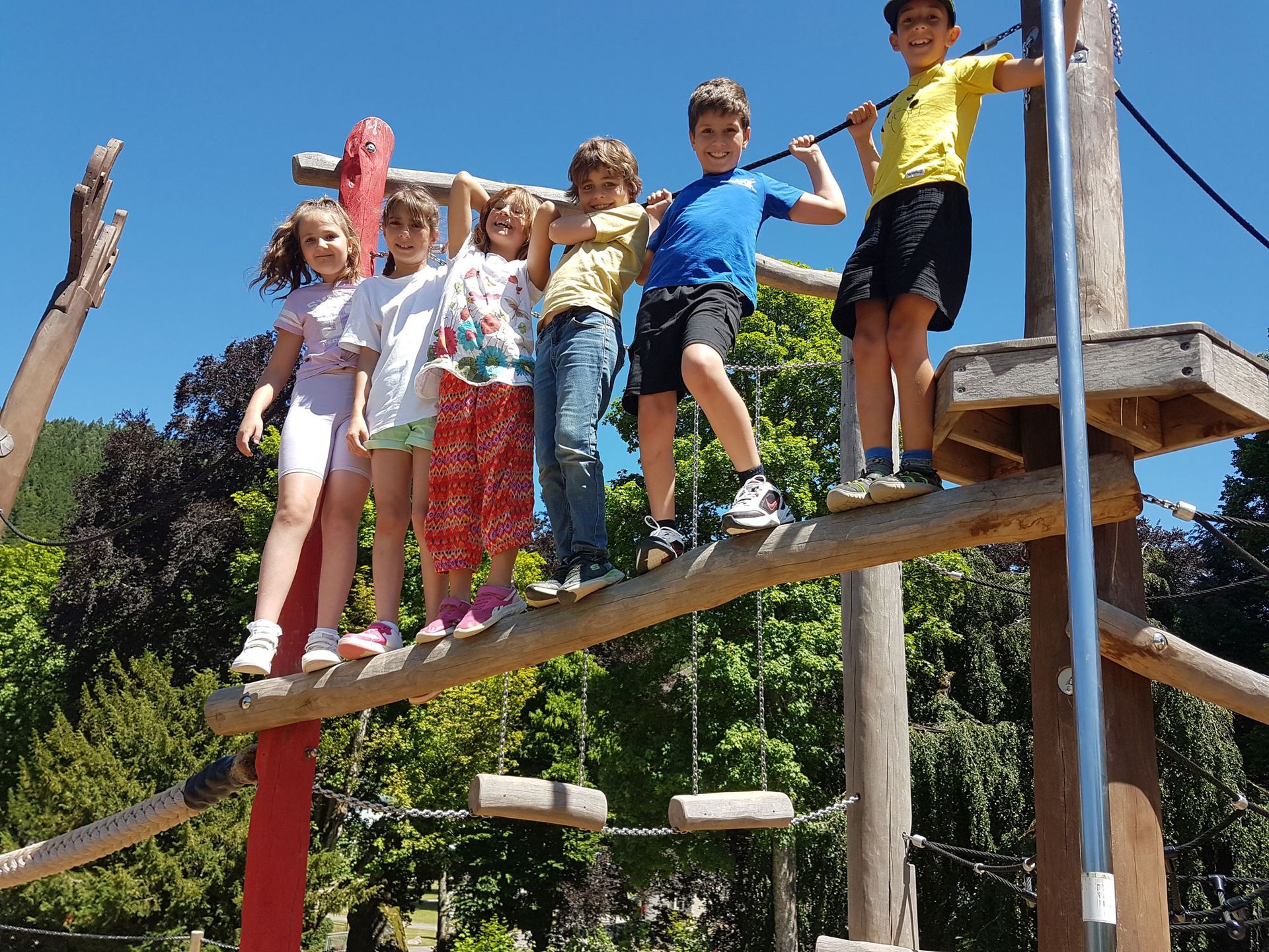 Kinder auf einem hölzernen Spielplatzgerüst, lächelnd und balancierend. Blauer Himmel, Grün im Hintergrund.