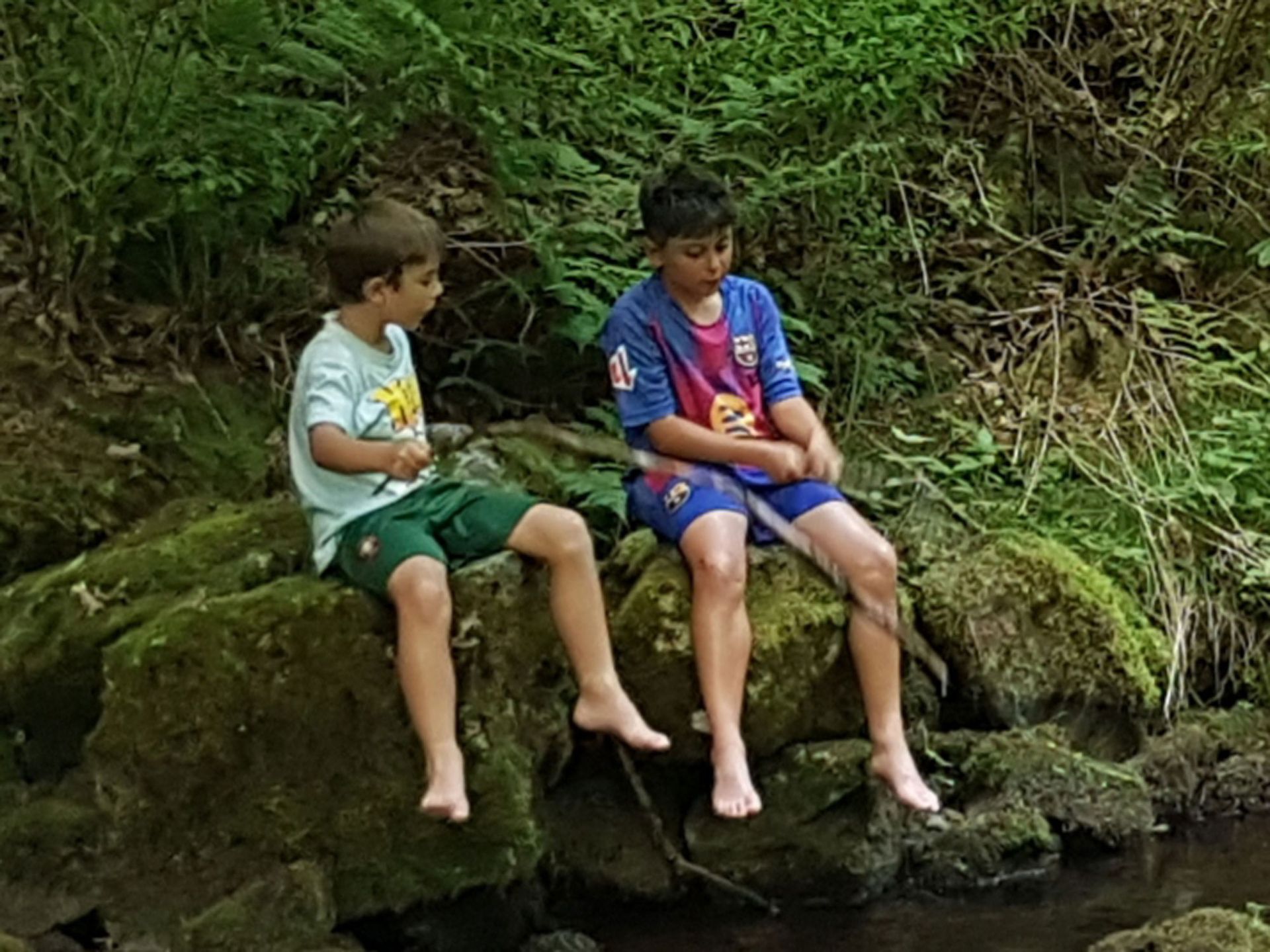 Zwei barfüßige Jungen sitzen auf moosbewachsenen Steinen an einem Bach und spielen mit Stöcken, umgeben von grünem Laub.