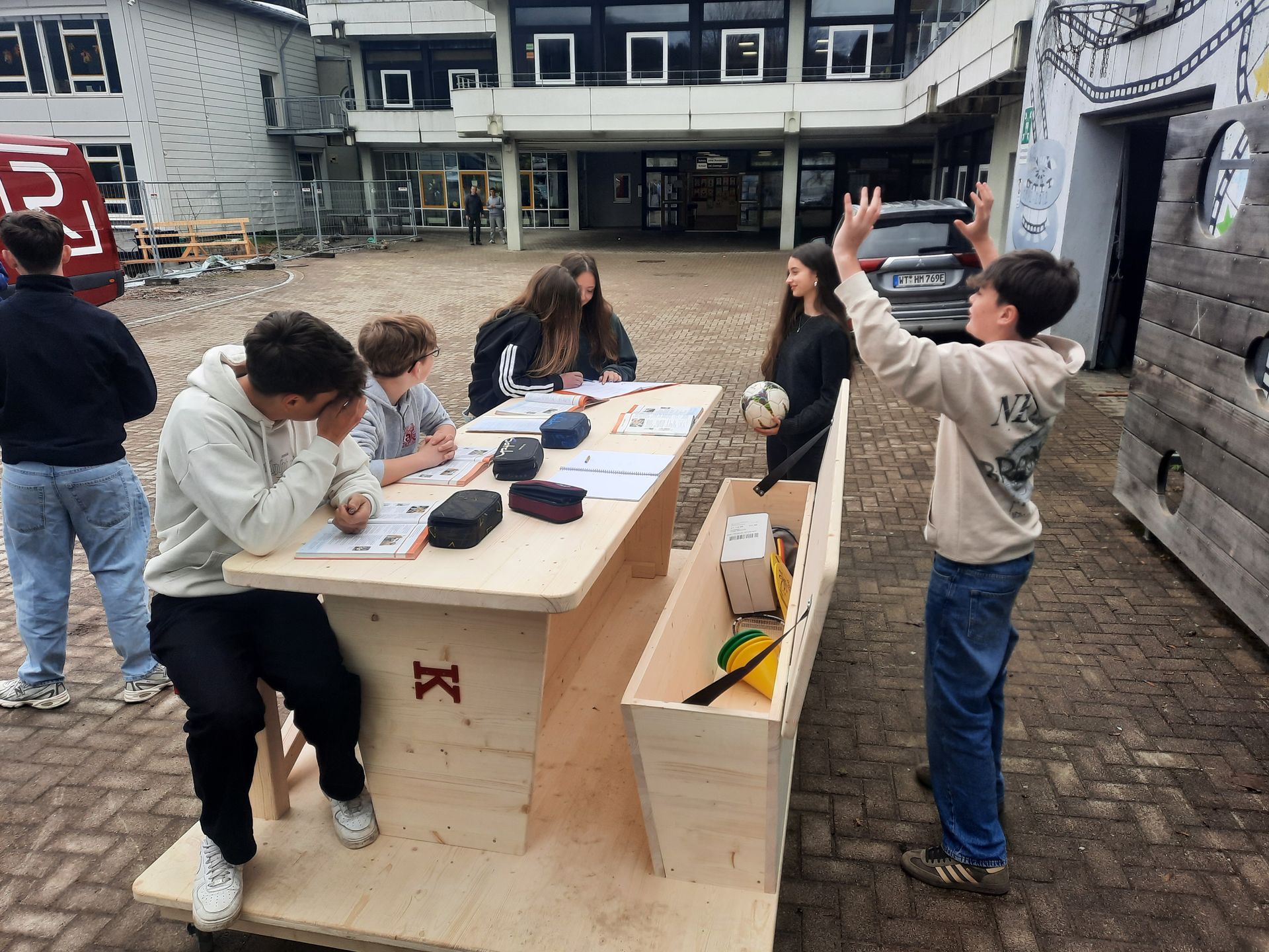 Die Leute versammelten sich draußen um einen Holztisch; eine Person hob die Hand, während andere Dinge ordneten.