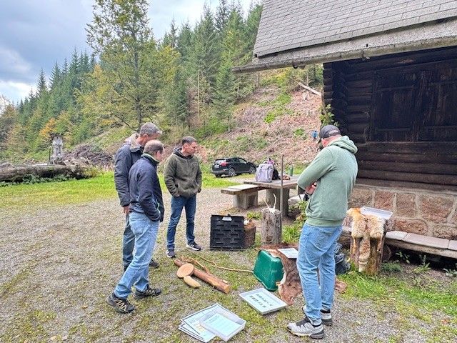 Vier Männer unterhalten sich im Freien in der Nähe einer Blockhütte über Gegenstände, die auf dem Boden liegen. Im Hintergrund sind grüne Bäume zu sehen.