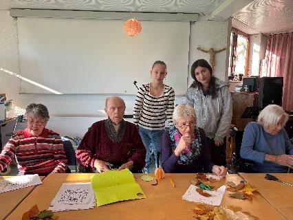 Eine Gruppe von Menschen bastelt an einem Tisch in einem Raum; zwei junge Frauen stehen hinter ihnen.