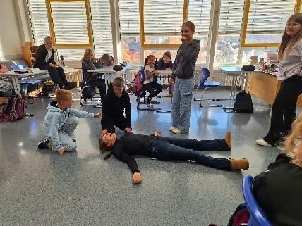 Kinder üben Erste Hilfe in einem Klassenzimmer, eines liegt auf dem Boden, die anderen beobachten.