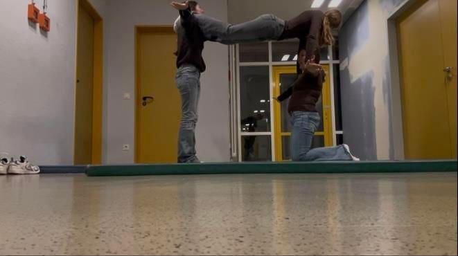 Drei Personen bilden in einem Innenraum eine akrobatische Menschenpyramide. Eine Person kniet, zwei stehen und halten eine dritte.