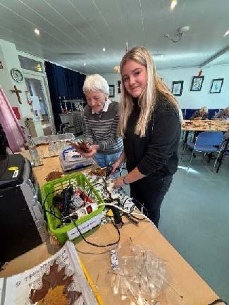 Zwei Personen basteln an einem Tisch. Ein blondes Mädchen lächelt, eine ältere Frau blickt auf die Materialien. Innenaufnahme.