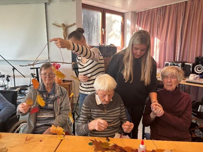 Menschen basteln mit Blättern; zwei Helfer unterstützen sie. Drinnen, bei Sonnenschein.