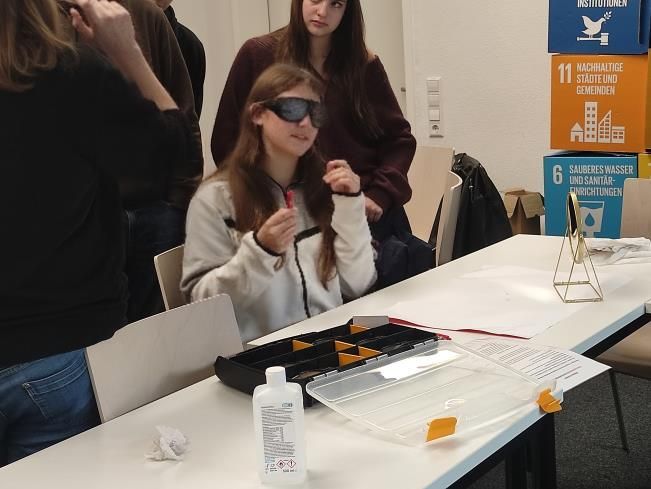 Ein Mädchen mit Sonnenbrille hält einen roten Gegenstand in der Hand und sitzt an einem Tisch. Im Hintergrund stehen weitere Personen an einer Tafel.