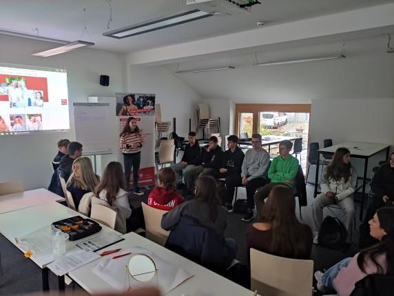Eine Präsentation in einem Klassenzimmer: Eine Frau spricht vor einer Gruppe von Schülern; im Hintergrund ist eine Projektionsfläche zu sehen.