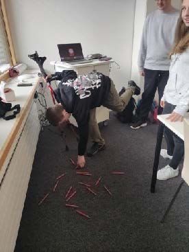 Menschen in einem Raum; einer ist vornübergebeugt und hebt rote Stifte vom Boden auf, zwei andere beobachten ihn, ein Laptop steht auf dem Schreibtisch.