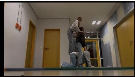 Drei Personen bilden eine Menschenpyramide in einem Flur mit gelben Türen und blauen Wänden.