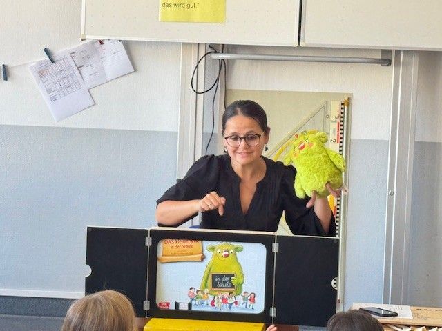 Eine Lehrerin führt mit einer Handpuppe vor einer Schulklasse ein Theaterstück auf. Auf der Puppenbühne ist eine Zeichnung eines grünen Monsters zu sehen.