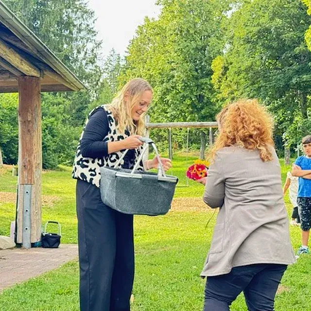 Eine Frau mit einem Korb nimmt in einem Park Blumen von einer anderen Frau entgegen.