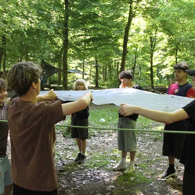 Eine Gruppe von Menschen hält ein weißes Laken im Freien in einem Waldgebiet.