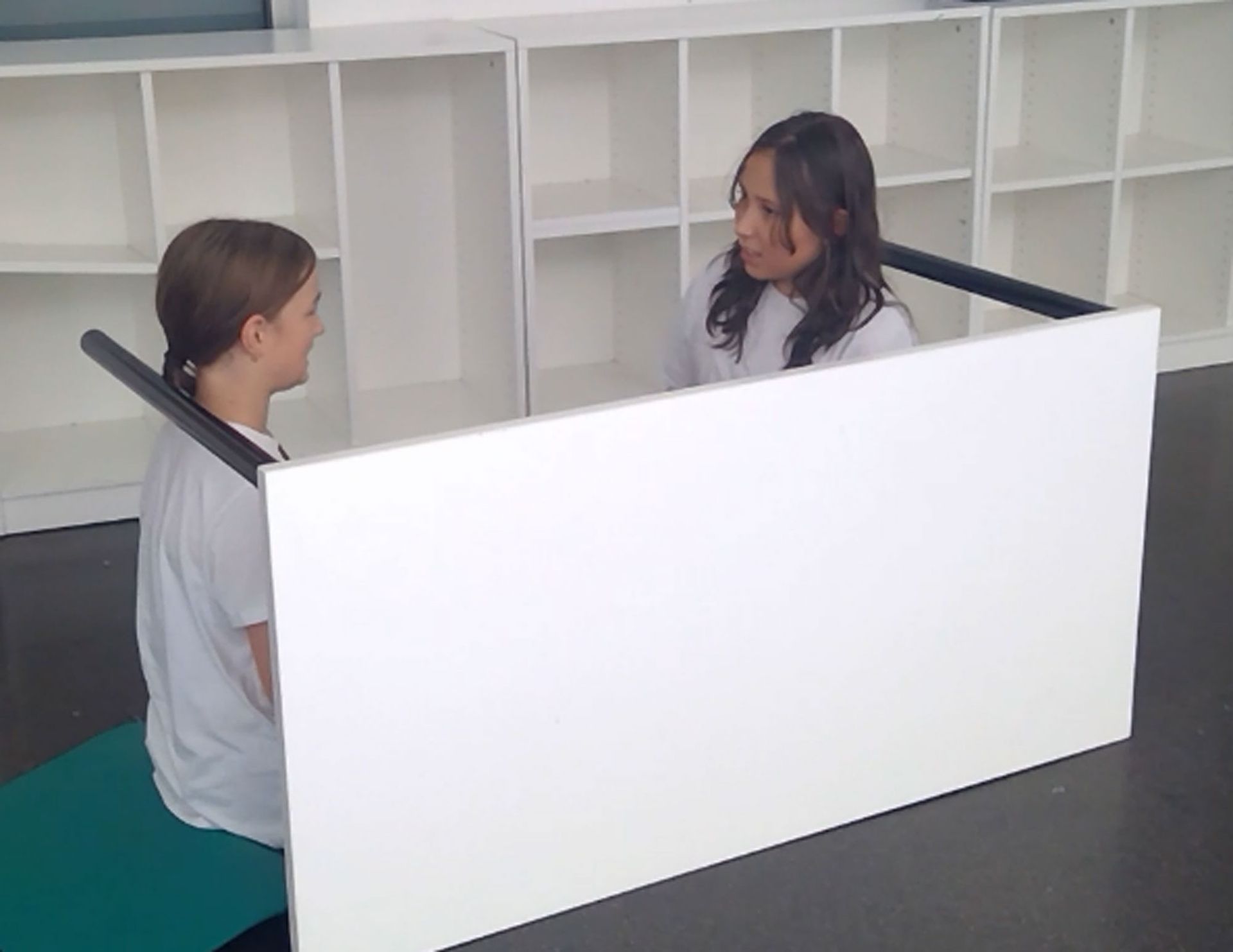 Zwei Personen sitzen sich gegenüber, getrennt durch eine weiße Trennwand, und unterhalten sich.