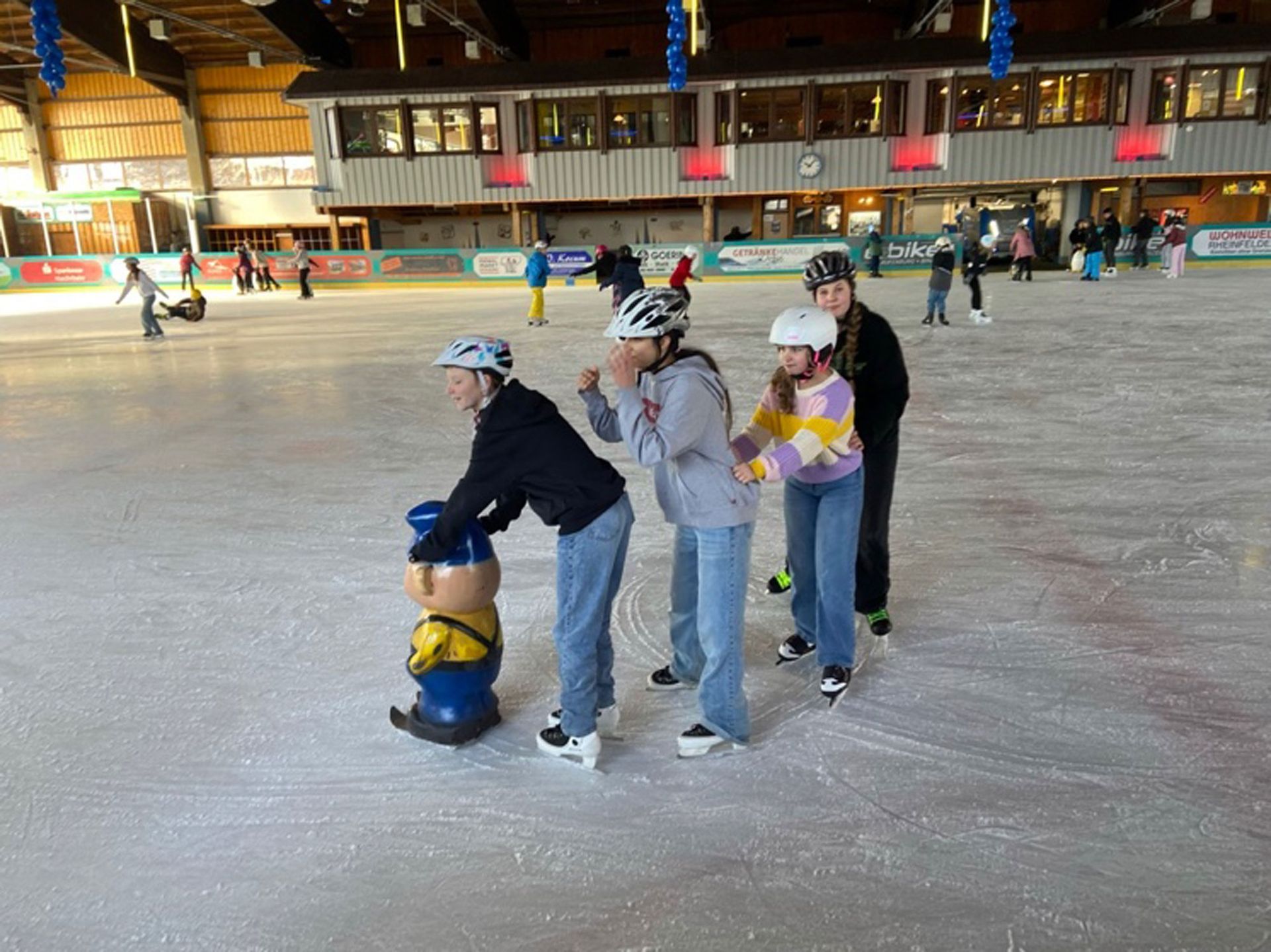 Vier Personen laufen in einer Halle Schlittschuh und benutzen dabei eine pinguinförmige Schlittschuhhilfe.