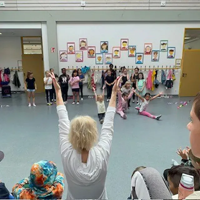 Eine Gruppe Kinder führt in einer Schulaula einen Tanz auf, angefeuert von einem Erwachsenen, der vorne steht.