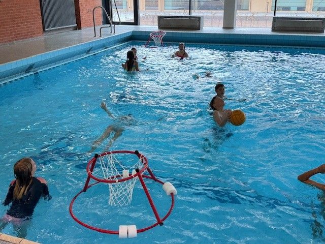 Kinder spielen in einem Schwimmbecken, einige mit einem Basketball. Ein Wasserbasketballkorb ist zu sehen.