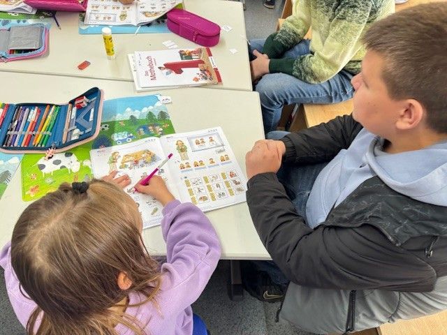 Zwei Kinder sitzen an einem Schreibtisch und erledigen ihre Schularbeiten. Ein Kind schreibt in ein Heft, das andere schaut zu.