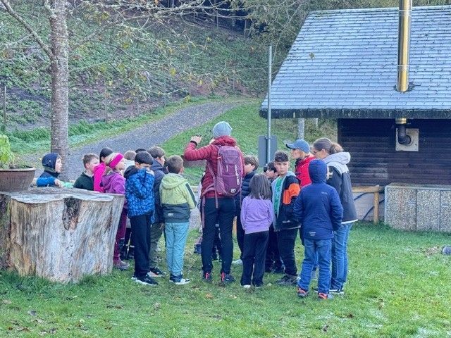Eine Gruppe Kinder hört im Freien vor einem kleinen Gebäude einer Person mit Rucksack zu.