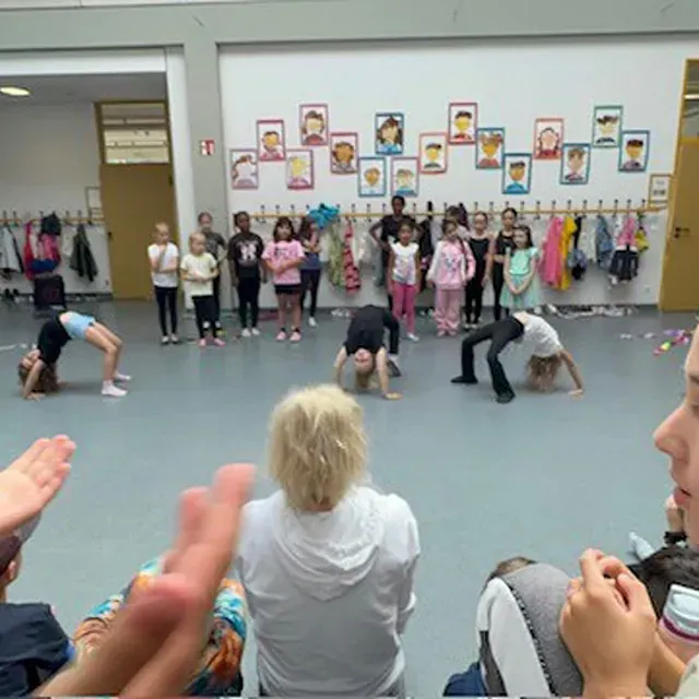 Kinder führen in einer Schulturnhalle Gymnastikübungen vor, während Zuschauer zusehen und applaudieren.