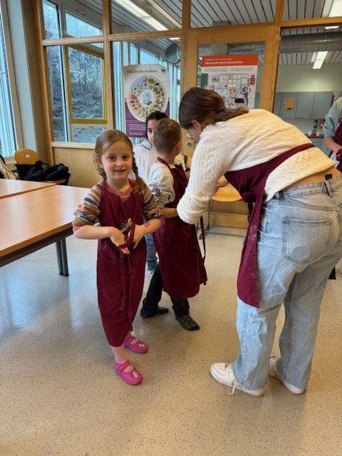 Mädchen in bordeauxroter Schürze lächelt, Lehrerin bindet ihr die Schürze um. Klassenzimmerszene, helle und neutrale Farbtöne.