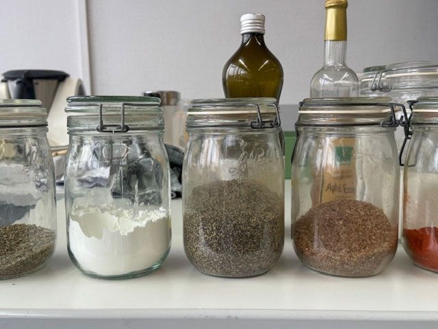 Auf einer Theke stehen mit Gewürzen und Zutaten gefüllte Glasgefäße, im Hintergrund sind Flaschen zu sehen.