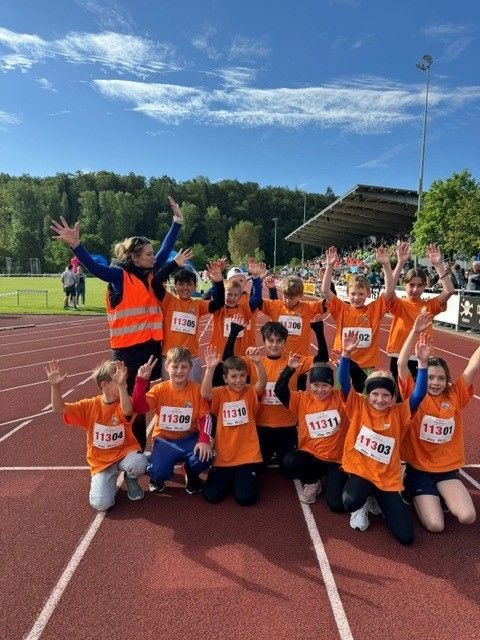 Children in orange race bibs posing on a track with medals and raised arms under a sunny sky