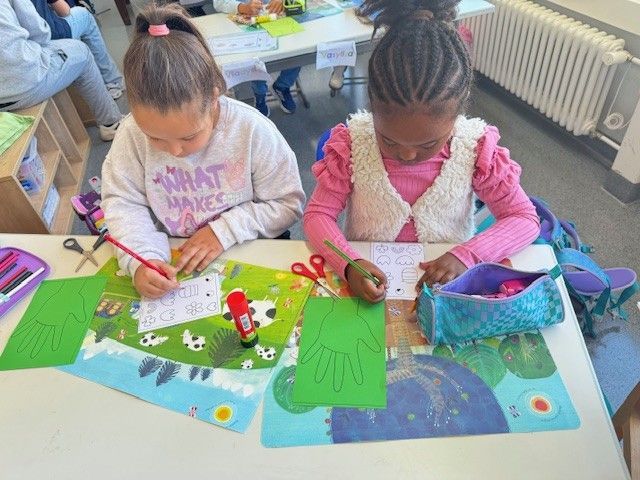 Zwei Kinder sitzen an einem Tisch und schreiben auf Papier. Sie basteln mit grünen Ausschnitten und anderen Materialien.